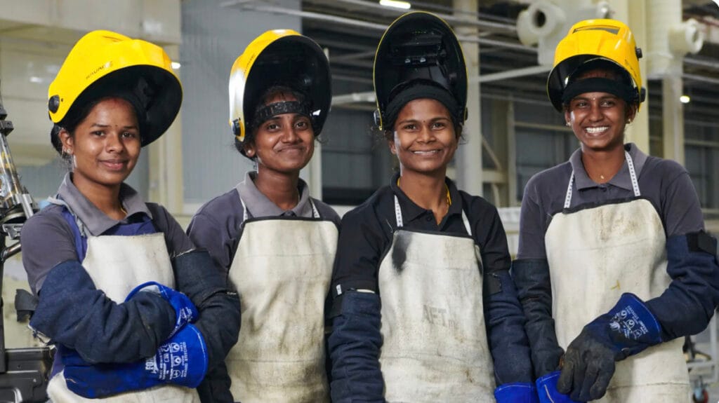 Four women wearing welding helmets, gloves, and protective aprons smiling at a factory worksite.