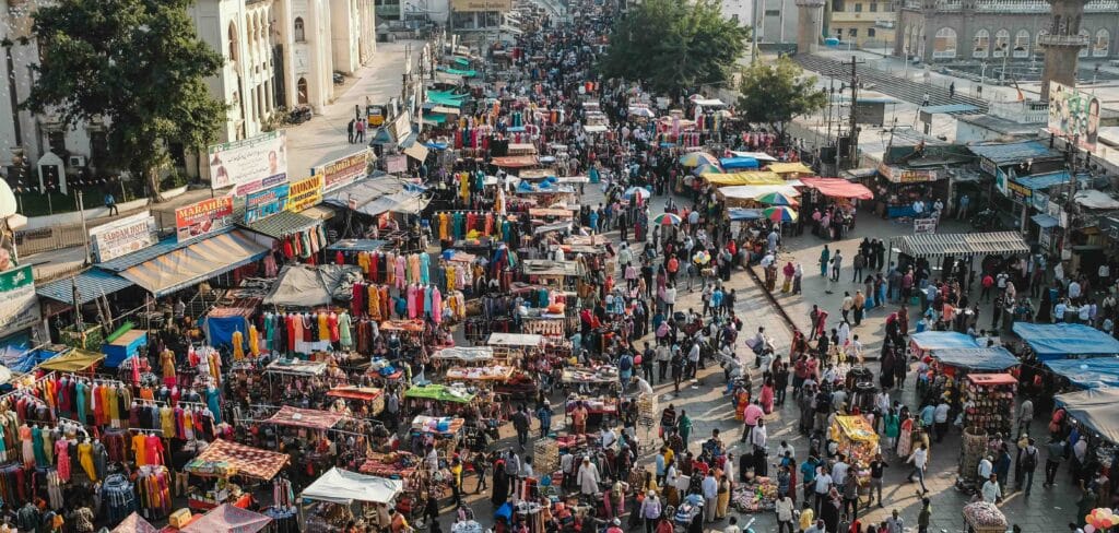 Aerial view of a crowded street market lined with colorful clothing stalls and dense pedestrian traffic.