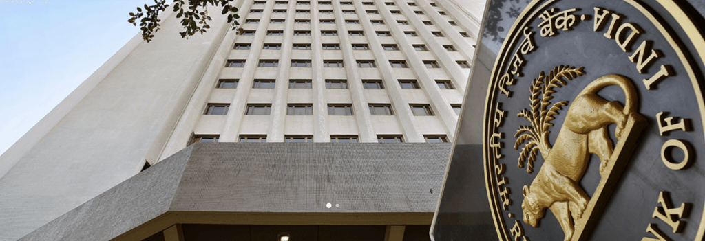 A low-angle, wide-shot of the Reserve Bank of India (RBI) headquarters in Mumbai, featuring the tall, white multi-story office building against a clear blue sky. In the foreground on the right, the gold-embossed RBI seal—depicting a tiger beneath a palm tree—is prominently displayed on a dark, polished plaque.
